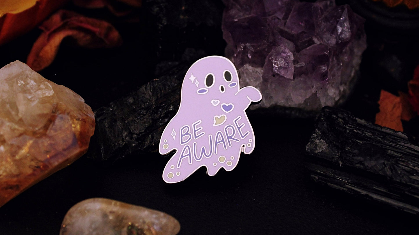 Be Aware Non-binary Pin- Silver plated enamel pin of a purple ghost with one hand sticking out and slightly bent, with the words "Be Aware" on it's body. The ghost also has black eyes and mouth, a white, purple, and yellow heart, yellow dots, and white stars, on it's body, based on the Non-binary flag colours. Original design by GRRRL Spells.