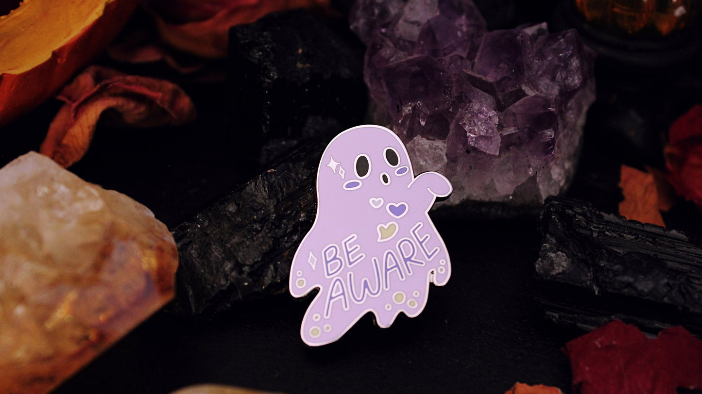 Be Aware Non-binary Pin- Silver plated enamel pin of a purple ghost with one hand sticking out and slightly bent, with the words "Be Aware" on it's body. The ghost also has black eyes and mouth, a white, purple, and yellow heart, yellow dots, and white stars, on it's body, based on the Non-binary flag colours. Original design by GRRRL Spells.