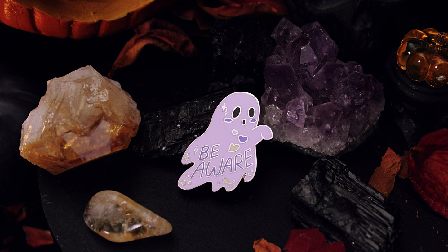 Be Aware Non-binary Pin- Silver plated enamel pin of a purple ghost with one hand sticking out and slightly bent, with the words "Be Aware" on it's body. The ghost also has black eyes and mouth, a white, purple, and yellow heart, yellow dots, and white stars, on it's body, based on the Non-binary flag colours. Original design by GRRRL Spells.