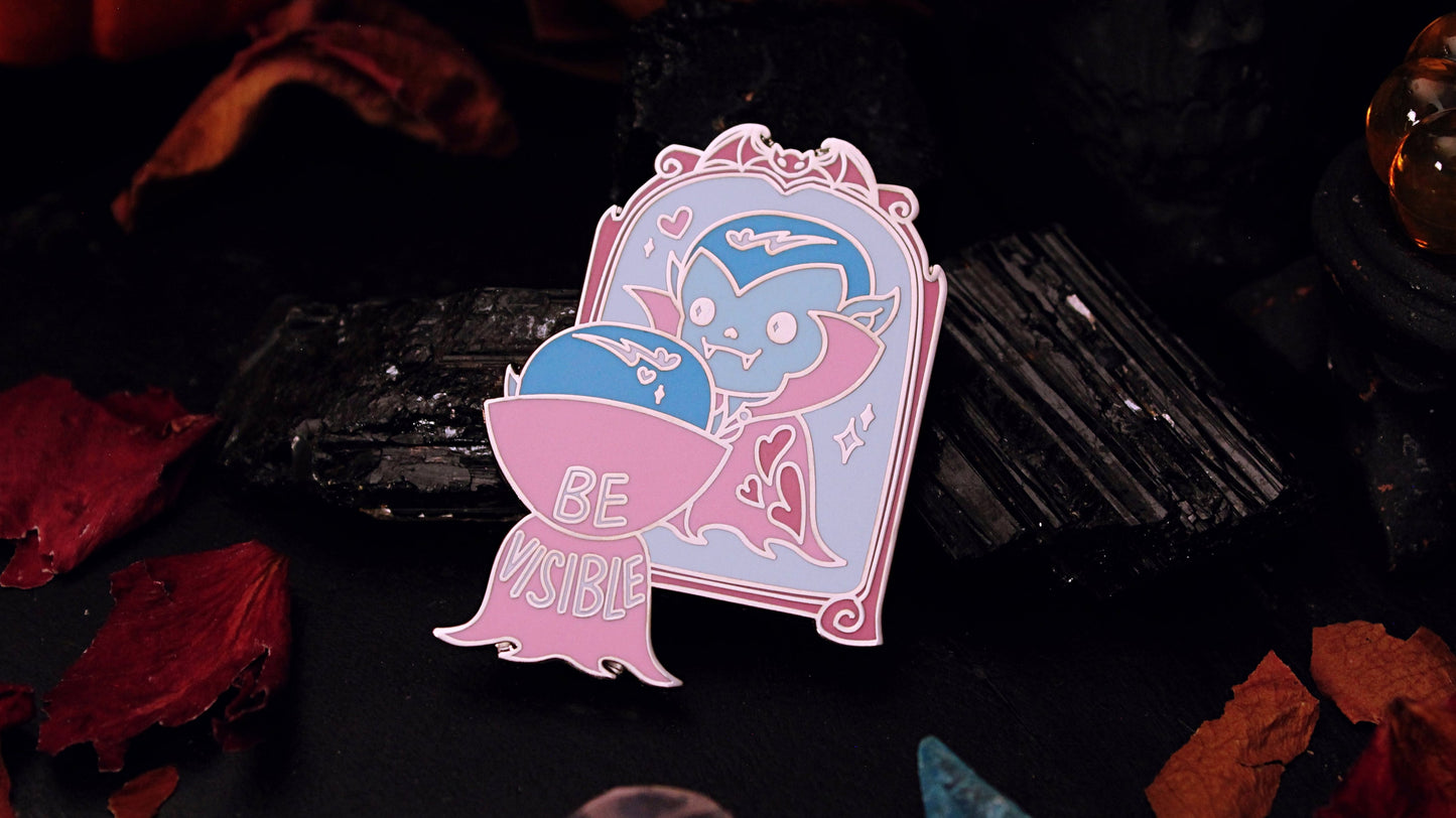 Be Visible Pin - Silver plated enamel pin of a cute vampire looking at their reflection in a mirror, in the trans flag colours of blue, pink, and white. The words Be Visible are seen on the back of their cape. Original design by GRRRL Spells.