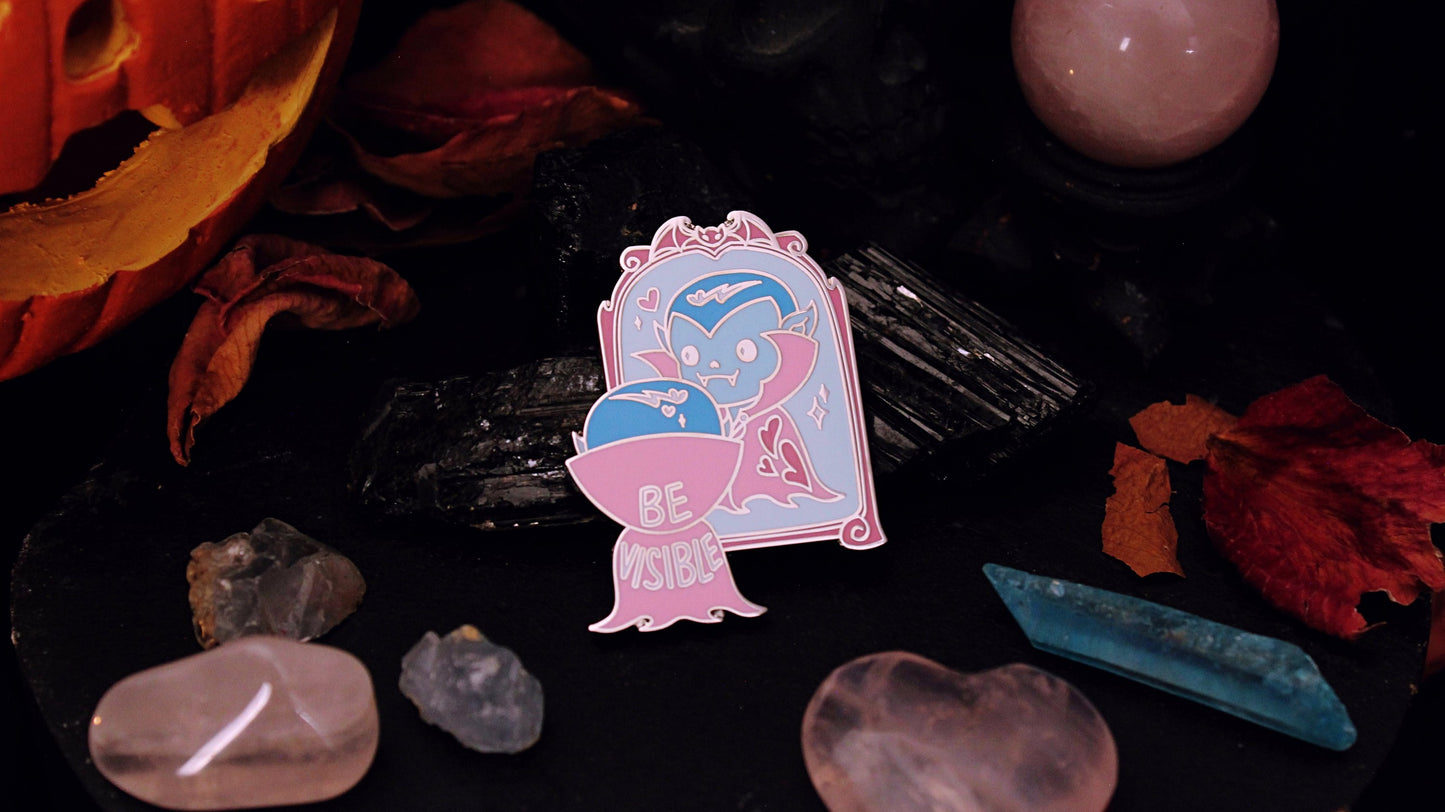 Be Visible Pin - Silver plated enamel pin of a cute vampire looking at their reflection in a mirror, in the trans flag colours of blue, pink, and white. The words Be Visible are seen on the back of their cape. Original design by GRRRL Spells.