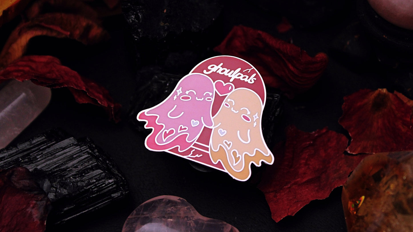 Ghoulpals Lesbian Pin - Silver plated enamel pin of two cute and smiling ghosts holding hands in front a tombstone that says "ghoulpals" with a heart in between them, in the colours of the lesbian flag which is orange, pink, and magenta. Original design by GRRRL Spells.