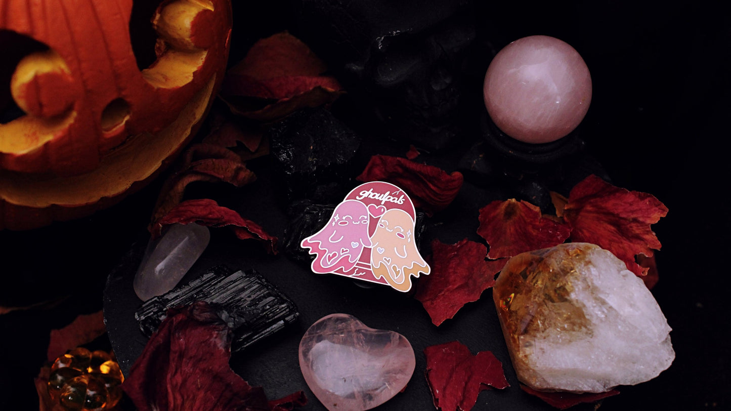 Ghoulpals Lesbian Pin - Silver plated enamel pin of two cute and smiling ghosts holding hands in front a tombstone that says "ghoulpals" with a heart in between them, in the colours of the lesbian flag which is orange, pink, and magenta. Original design by GRRRL Spells.