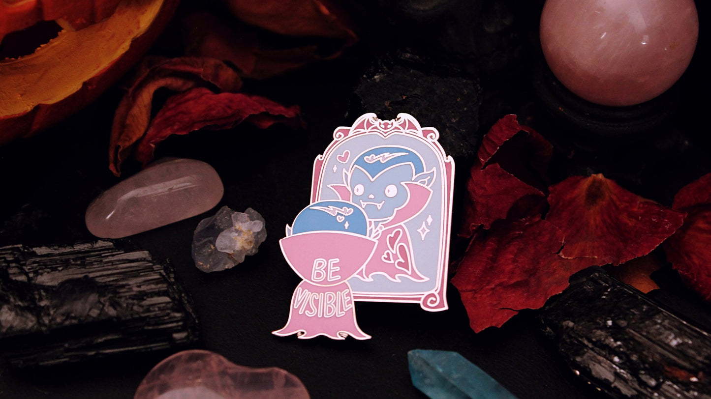Be Visible Pin - Silver plated enamel pin of a cute vampire looking at their reflection in a mirror, in the trans flag colours of blue, pink, and white. The words Be Visible are seen on the back of their cape. Original design by GRRRL Spells.