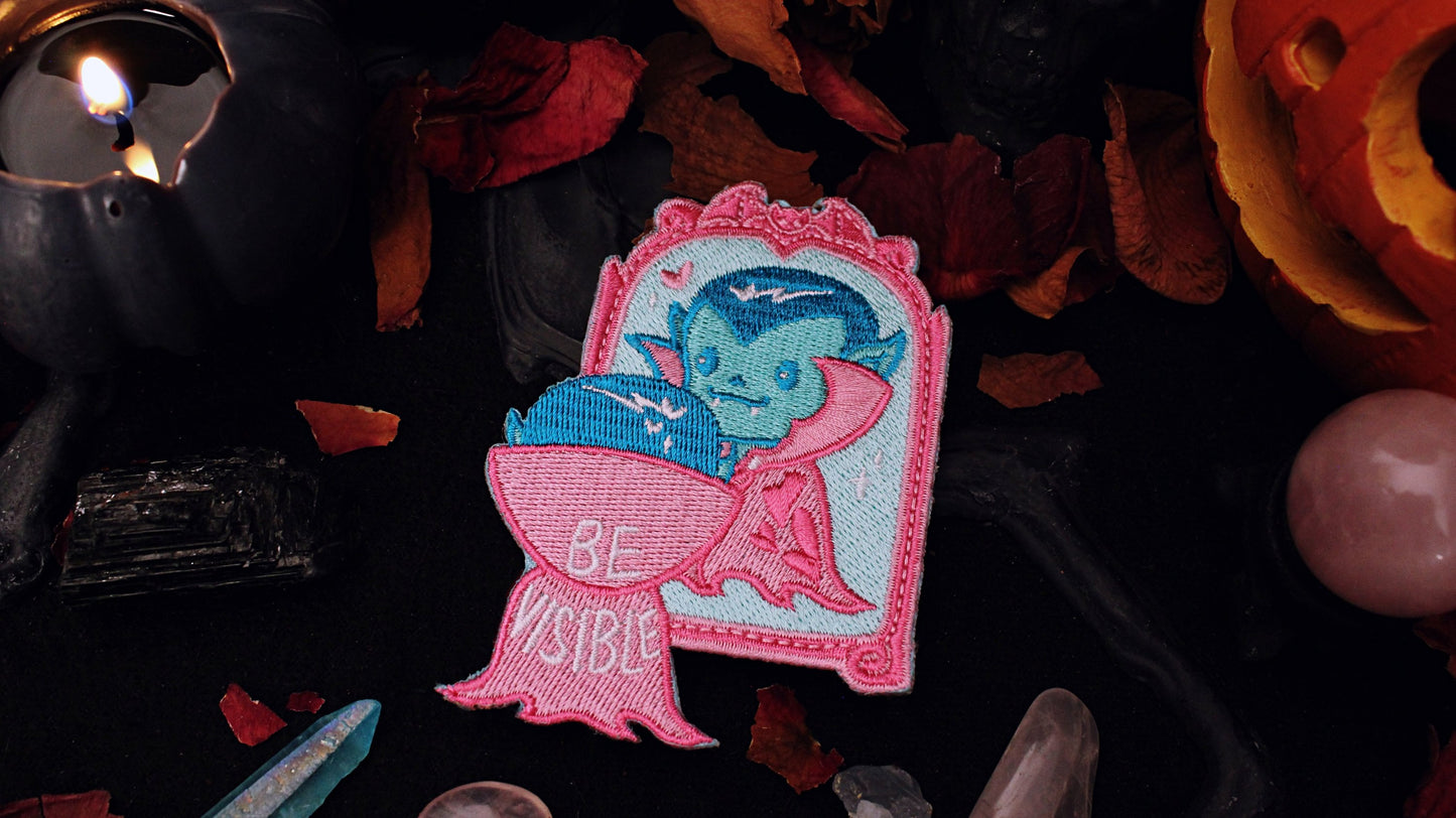 Be Visible Patch - Iron on embroidered patch of a cute vampire looking at their reflection in a mirror, in the trans flag colours of blue, pink, and white. The words Be Visible are seen on the back of their cape. Original design by GRRRL Spells.