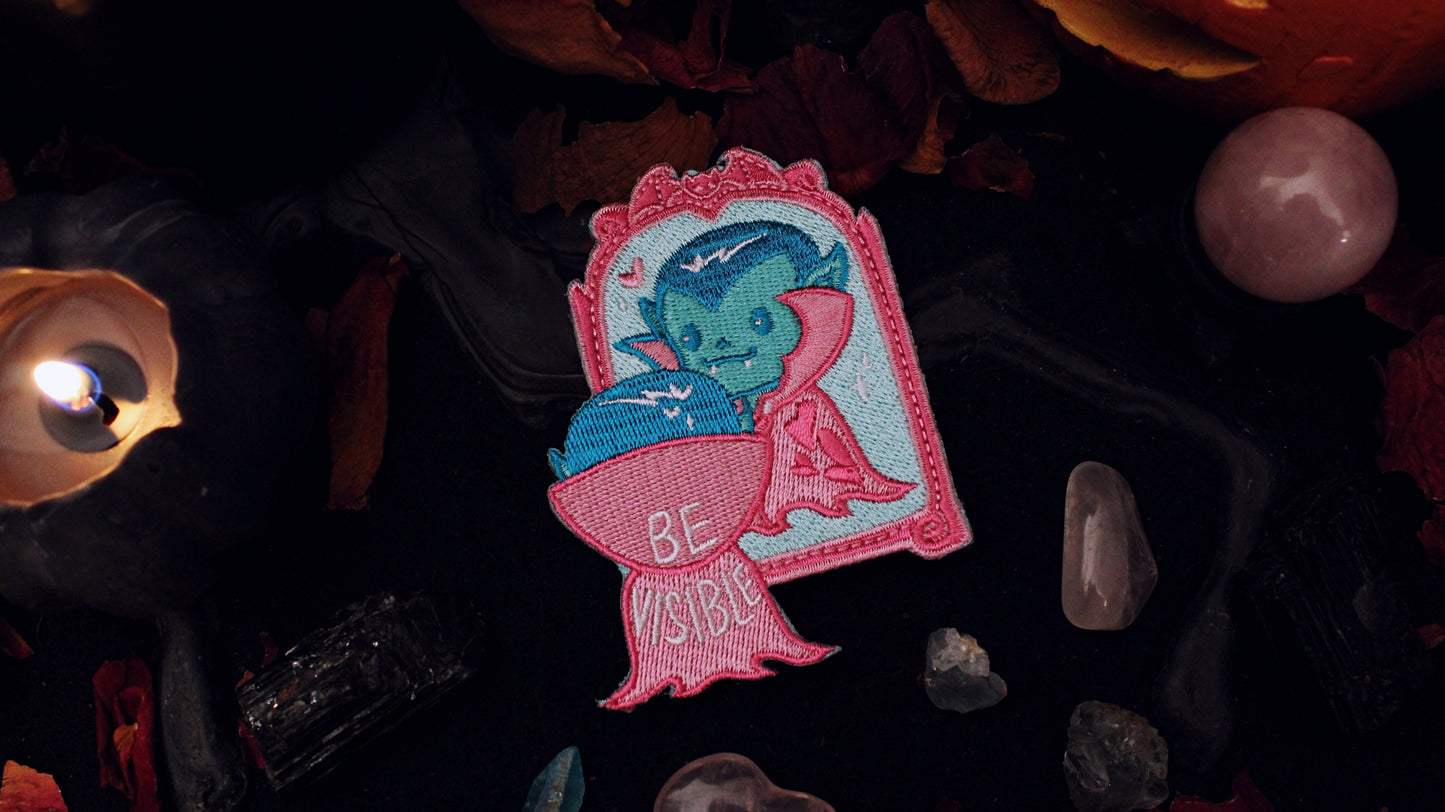 Be Visible Patch - Iron on embroidered patch of a cute vampire looking at their reflection in a mirror, in the trans flag colours of blue, pink, and white. The words Be Visible are seen on the back of their cape. Original design by GRRRL Spells.