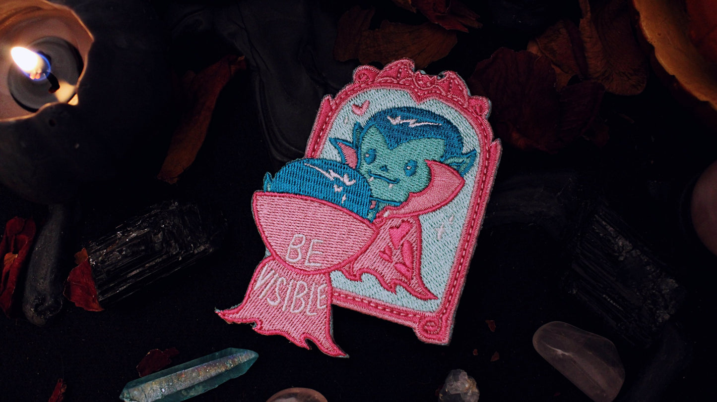Be Visible Patch - Iron on embroidered patch of a cute vampire looking at their reflection in a mirror, in the trans flag colours of blue, pink, and white. The words Be Visible are seen on the back of their cape. Original design by GRRRL Spells.