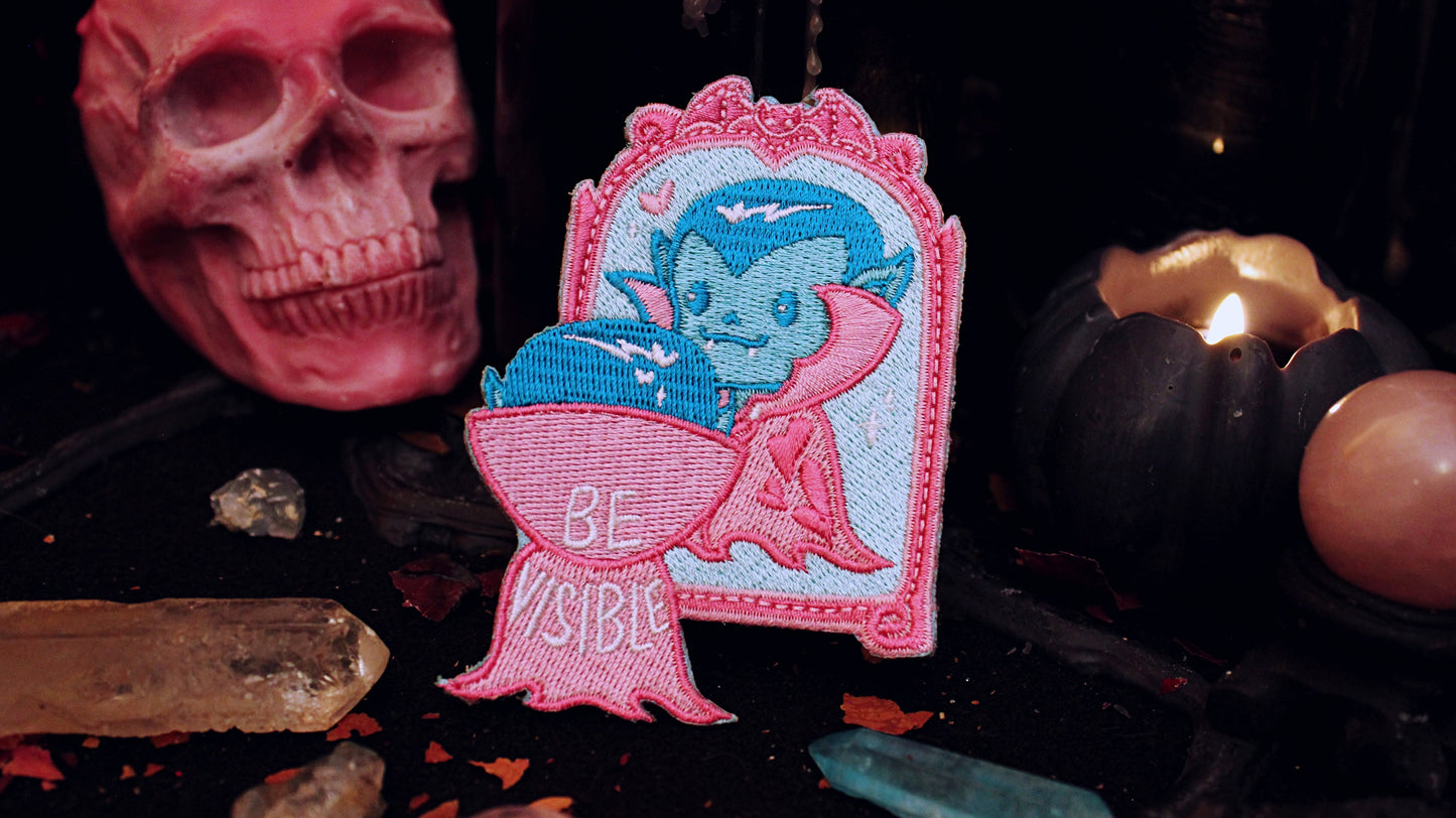 Be Visible Patch - Iron on embroidered patch of a cute vampire looking at their reflection in a mirror, in the trans flag colours of blue, pink, and white. The words Be Visible are seen on the back of their cape. Original design by GRRRL Spells.