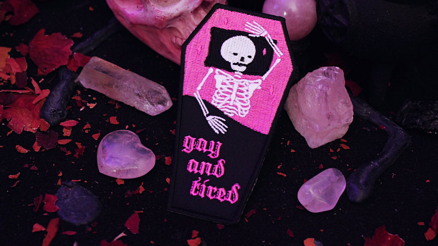 Iron on embroidered patch that is coffin shaped with black border and half pink on the top and half black on the bottom as background. Skeleton lays in the coffin bed exhausted as the words "gay and tired" in lowercase letters written on the bottom half of the coffin in gothic typography. Original design by GRRRL Spells.