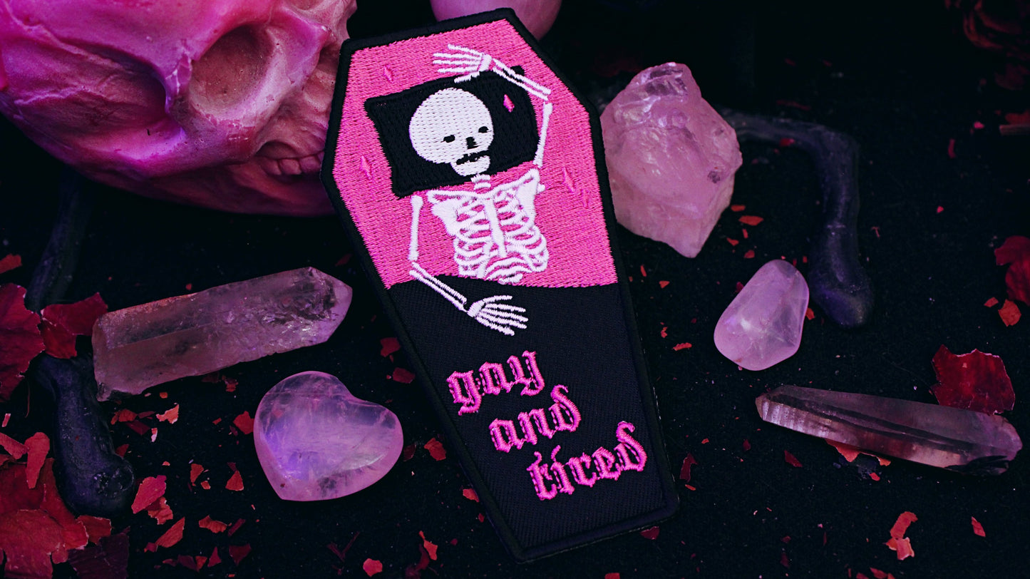 Iron on embroidered patch that is coffin shaped with black border and half pink on the top and half black on the bottom as background. Skeleton lays in the coffin bed exhausted as the words "gay and tired" in lowercase letters written on the bottom half of the coffin in gothic typography. Original design by GRRRL Spells.