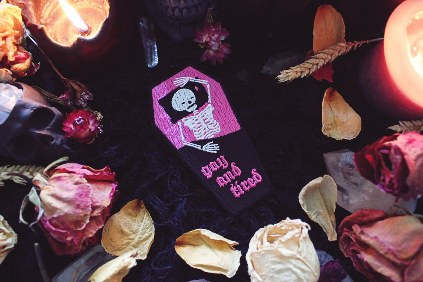 Iron on embroidered patch that is coffin shaped with black border and half pink on the top and half black on the bottom as background. Skeleton lays in the coffin bed exhausted as the words "gay and tired" in lowercase letters written on the bottom half of the coffin in gothic typography. Original design by GRRRL Spells.