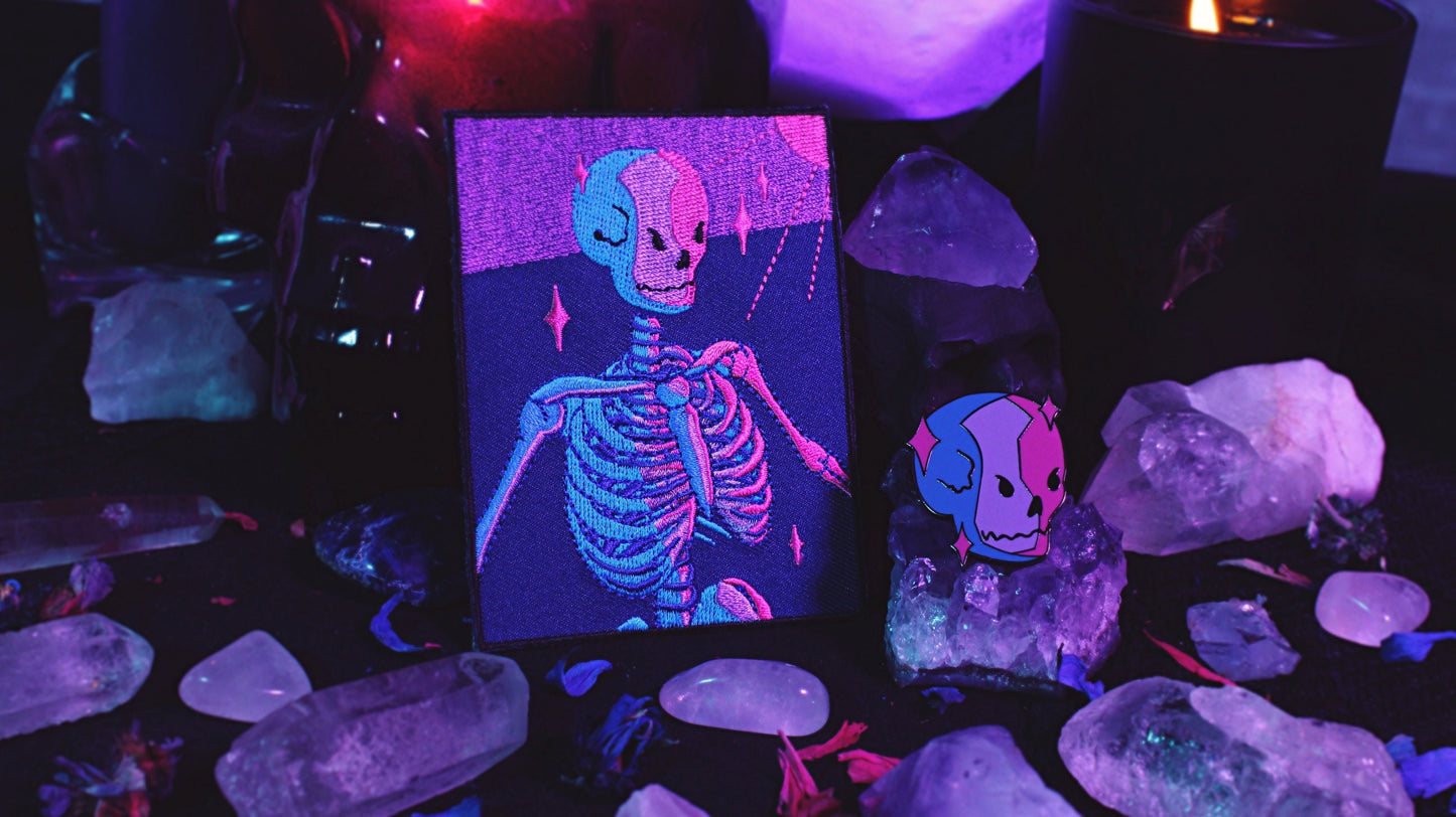 Bisexual Lighting Skeleton Patch - Iron on embroidered patch of a portrait of a skeleton in bisexual lighting with the colours blue, pink, and purple. There is a ray of pink lighting from the top right corner with glimmering pink sparkle around the skeleton. Original design by GRRRL Spells.