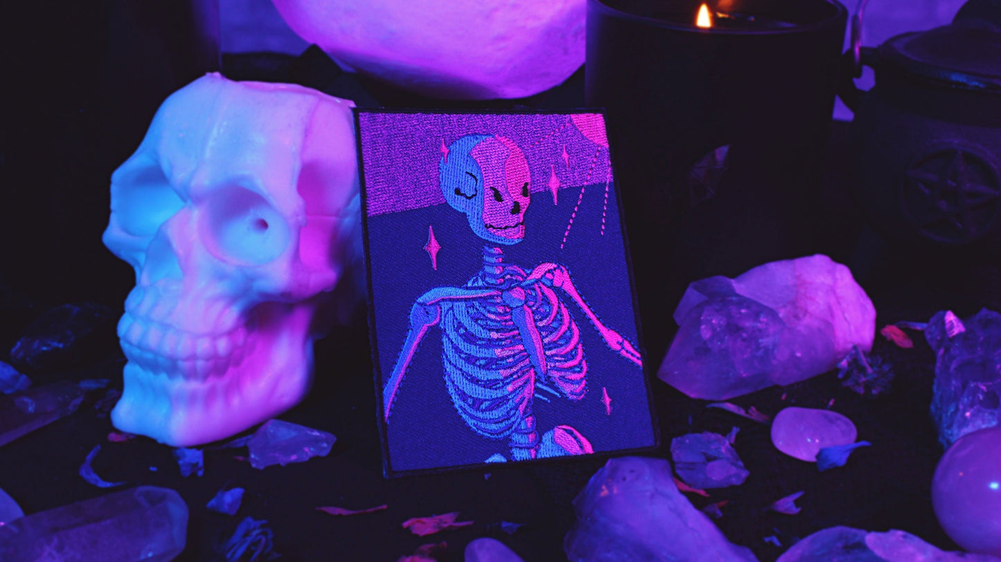 Bisexual Lighting Skeleton Patch - Iron on embroidered patch of a portrait of a skeleton in bisexual lighting with the colours blue, pink, and purple. There is a ray of pink lighting from the top right corner with glimmering pink sparkle around the skeleton. Original design by GRRRL Spells.
