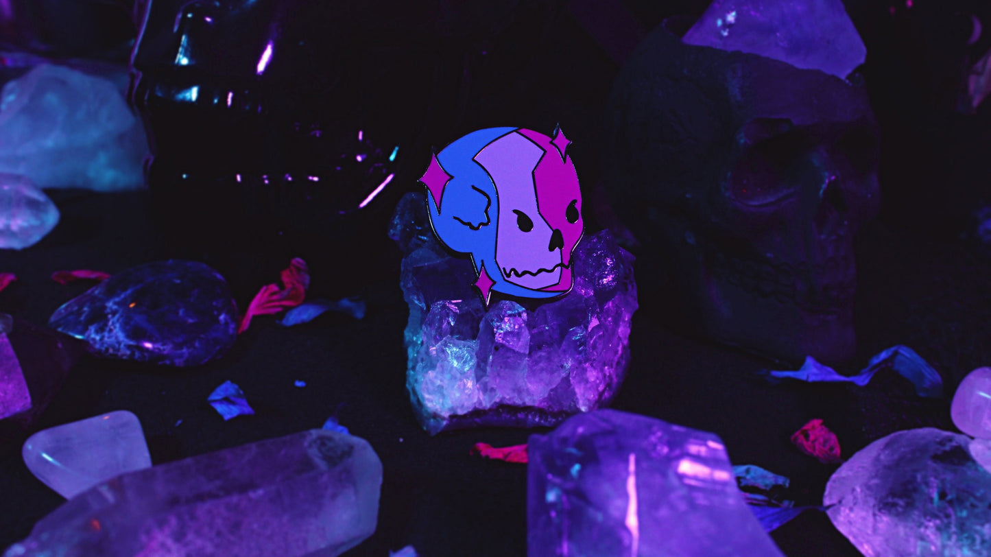 Bisexual Lighting Skeleton Pin - Black nickel plated enamel pin of a skeleton in bisexual lighting with the colours blue, pink, and purple with pink sparkles around the skull. Original design by GRRRL Spells.