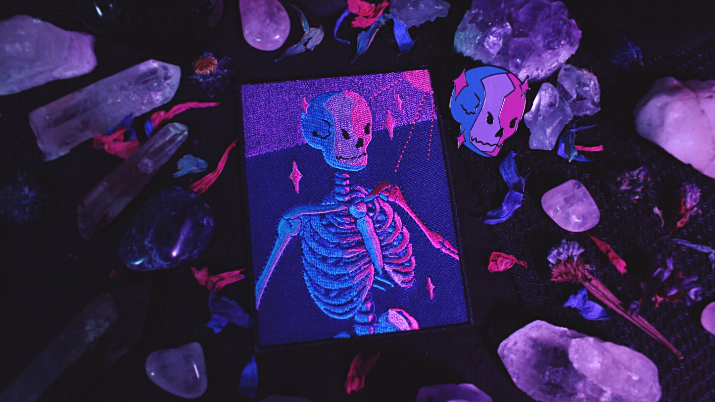 Bisexual Lighting Skeleton Patch - Iron on embroidered patch of a portrait of a skeleton in bisexual lighting with the colours blue, pink, and purple. There is a ray of pink lighting from the top right corner with glimmering pink sparkle around the skeleton. Original design by GRRRL Spells.