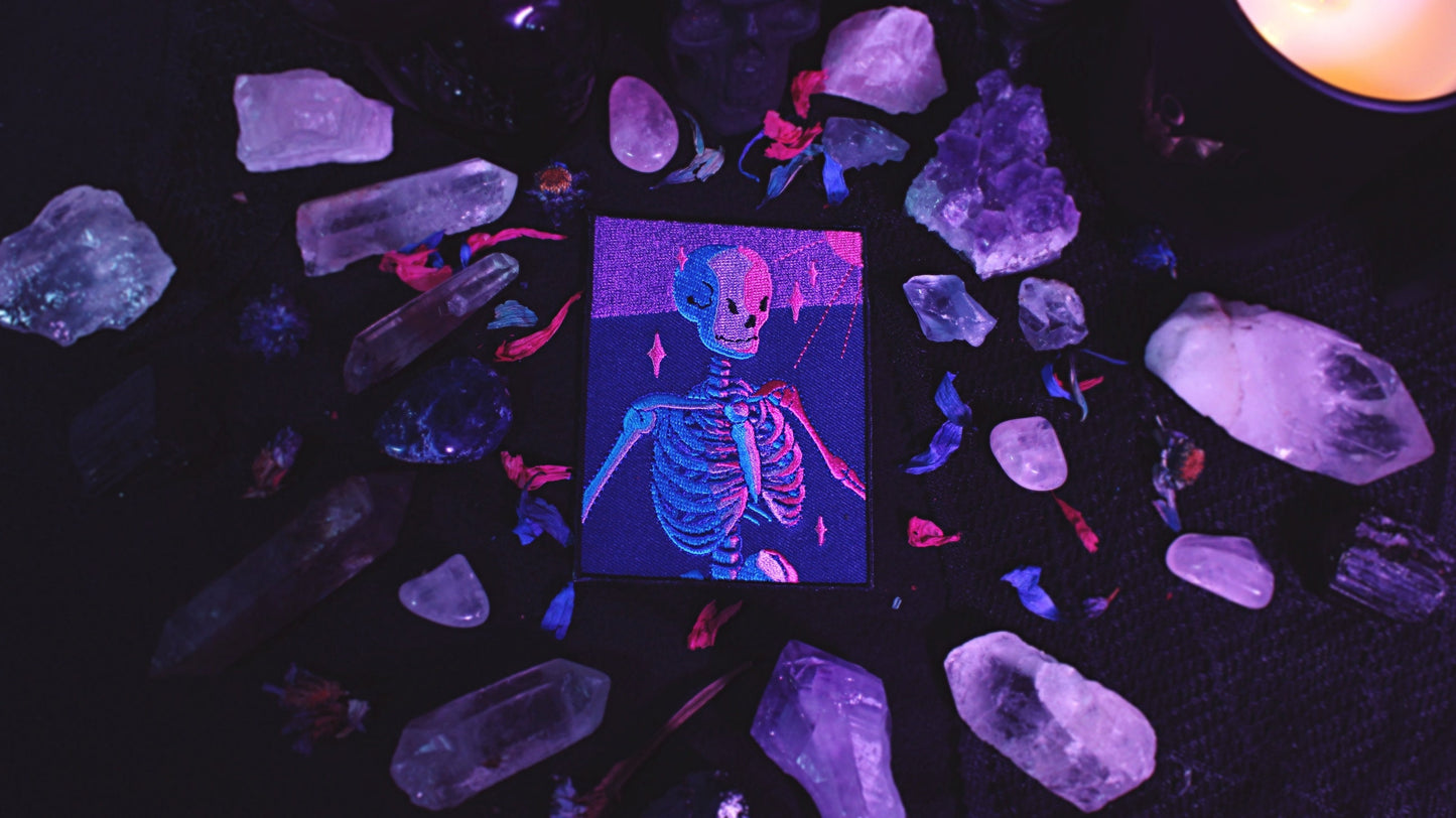 Bisexual Lighting Skeleton Patch - Iron on embroidered patch of a portrait of a skeleton in bisexual lighting with the colours blue, pink, and purple. There is a ray of pink lighting from the top right corner with glimmering pink sparkle around the skeleton. Original design by GRRRL Spells.