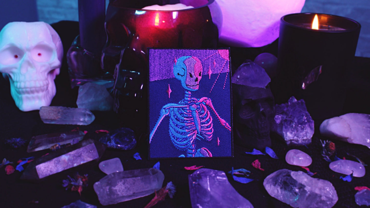 Bisexual Lighting Skeleton Patch - Iron on embroidered patch of a portrait of a skeleton in bisexual lighting with the colours blue, pink, and purple. There is a ray of pink lighting from the top right corner with glimmering pink sparkle around the skeleton. Original design by GRRRL Spells.
