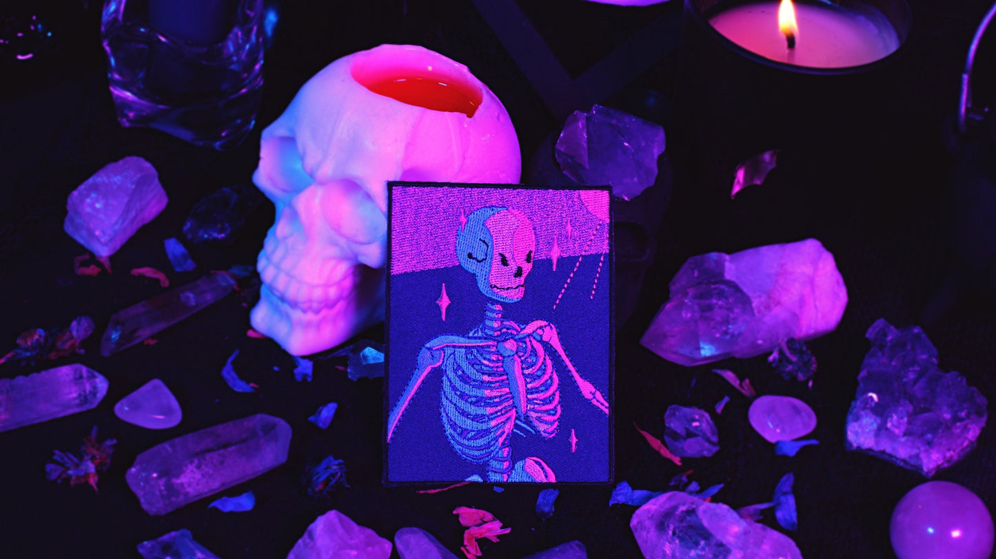Bisexual Lighting Skeleton Patch - Iron on embroidered patch of a portrait of a skeleton in bisexual lighting with the colours blue, pink, and purple. There is a ray of pink lighting from the top right corner with glimmering pink sparkle around the skeleton. Original design by GRRRL Spells.