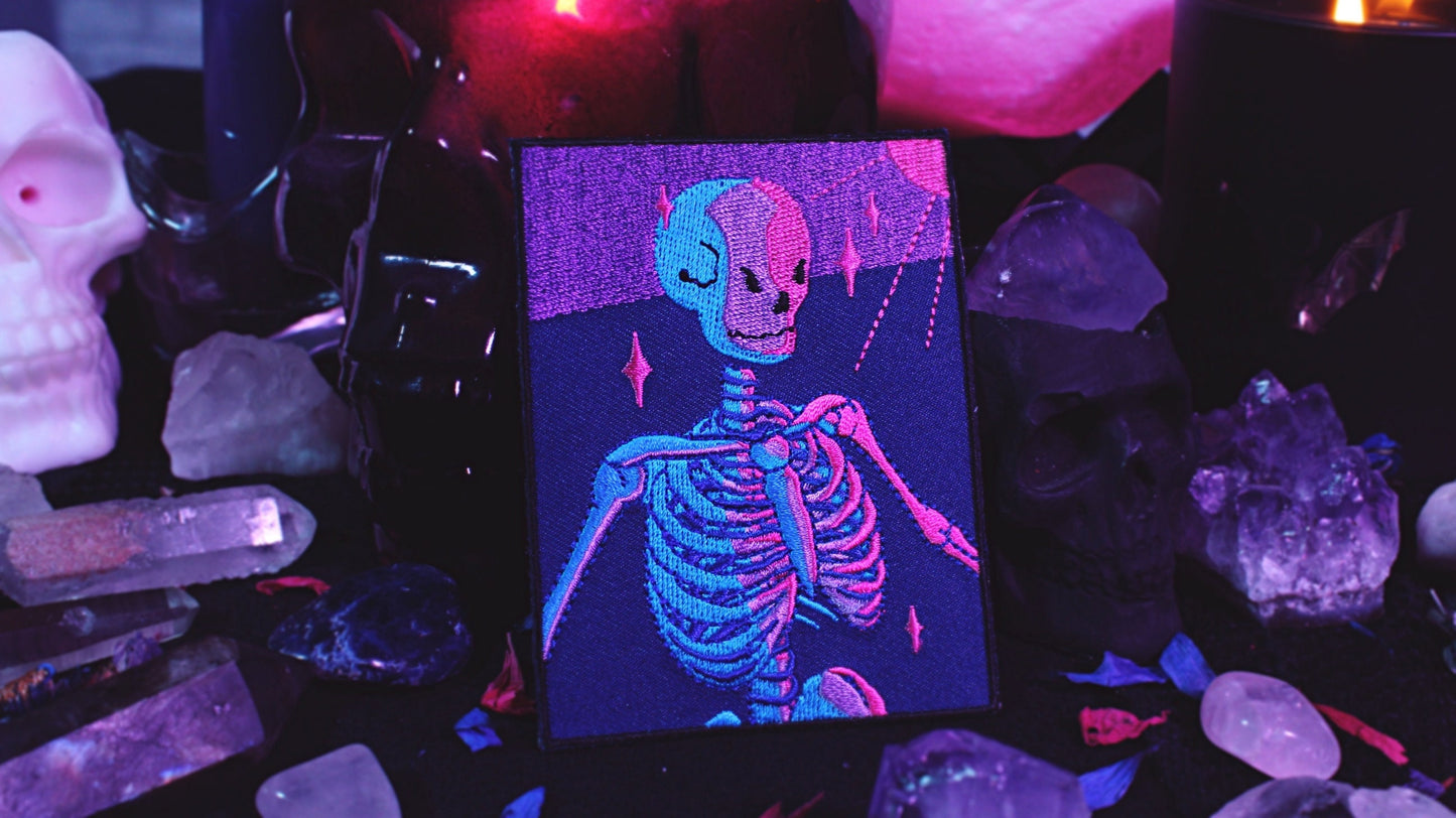 Bisexual Lighting Skeleton Patch - Iron on embroidered patch of a portrait of a skeleton in bisexual lighting with the colours blue, pink, and purple. There is a ray of pink lighting from the top right corner with glimmering pink sparkle around the skeleton. Original design by GRRRL Spells.