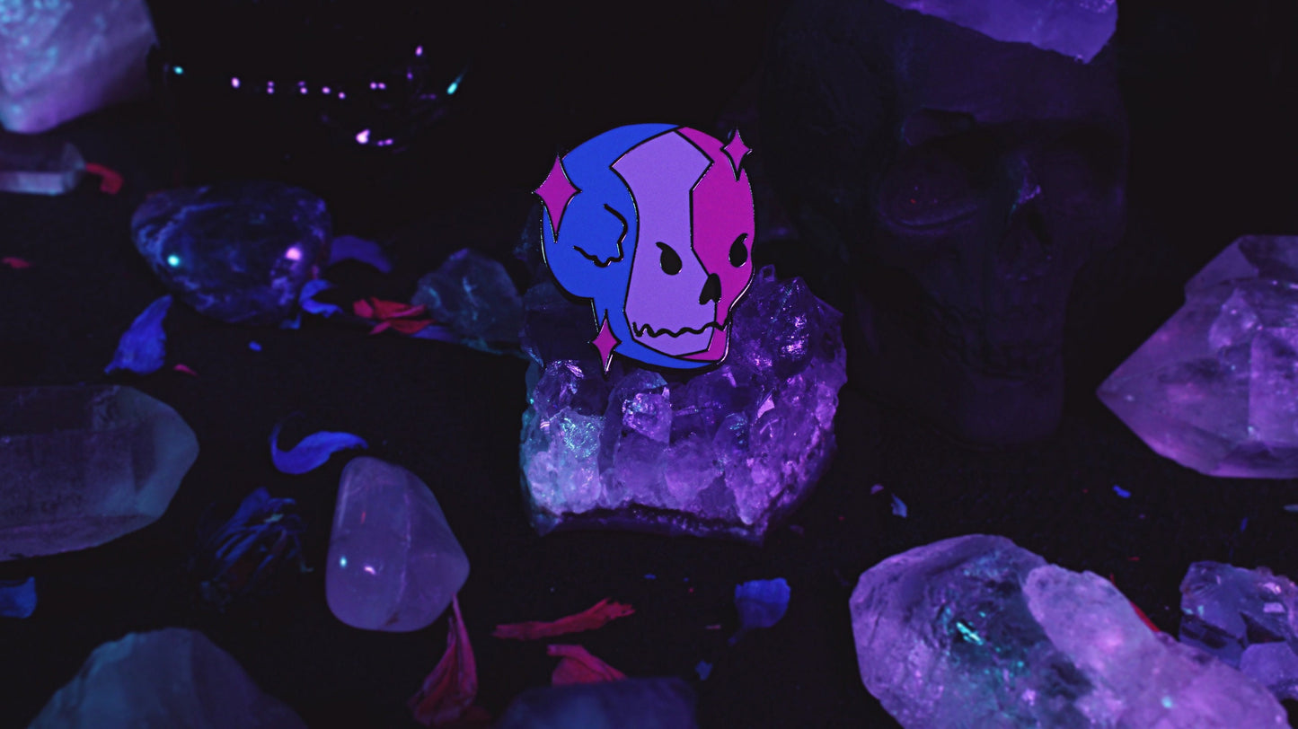 Bisexual Lighting Skeleton Pin - Black nickel plated enamel pin of a skeleton in bisexual lighting with the colours blue, pink, and purple with pink sparkles around the skull. Original design by GRRRL Spells.