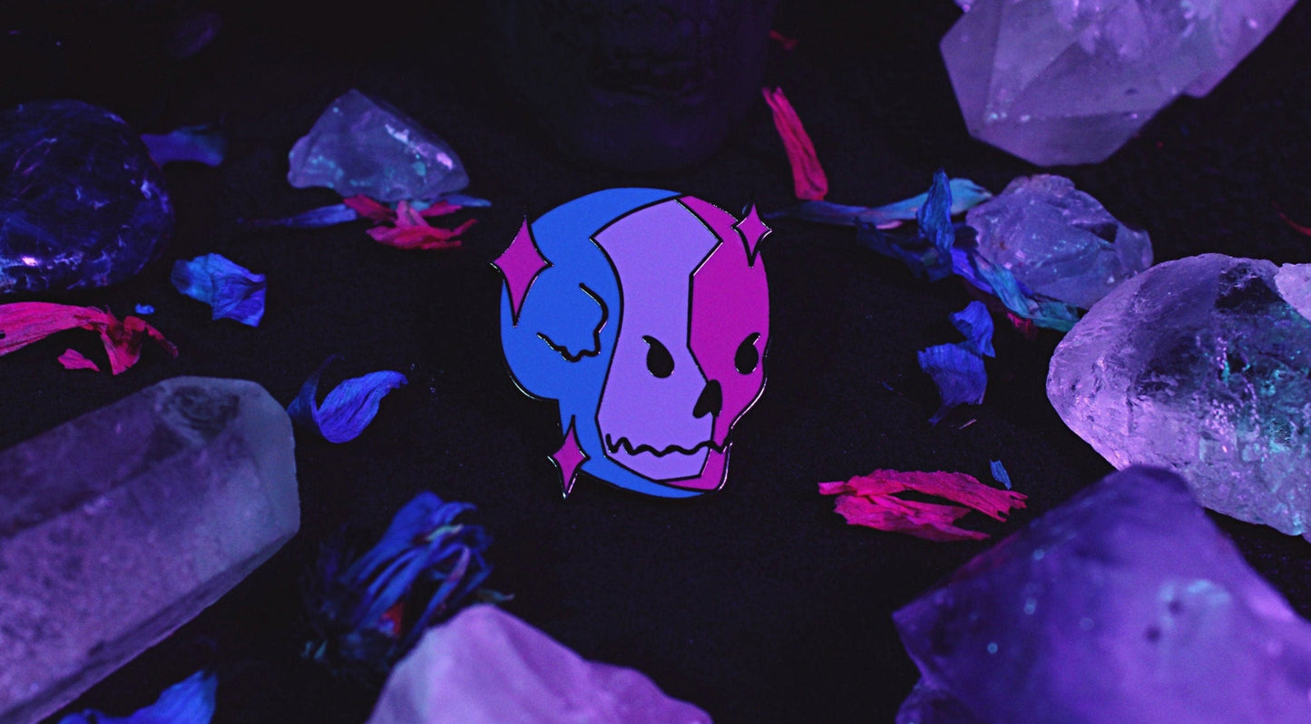Bisexual Lighting Skeleton Pin - Black nickel plated enamel pin of a skeleton in bisexual lighting with the colours blue, pink, and purple with pink sparkles around the skull. Original design by GRRRL Spells.