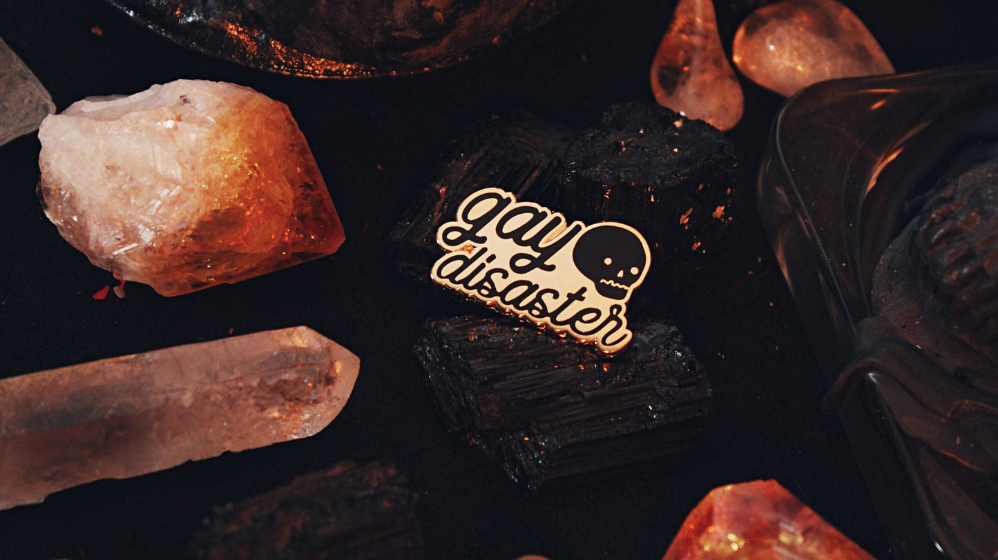 Gay Disaster Pin - Gold plated enamel pin with the words "gay disaster" written in black in lowercase in handwritten semi-cursive typography with a cute skull in the top right corner. Original design by GRRRL Spells.