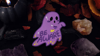 Be Aware Non-binary Patch - Iron on embroidered patch of a purple ghost with one hand sticking out and slightly bent, with the words "Be Aware" on it's body. The ghost also has black eyes and mouth, a white, purple, and yellow heart, yellow dots, and white stars, on it's body, based on the Non-binary flag colours. Original design by GRRRL Spells. 