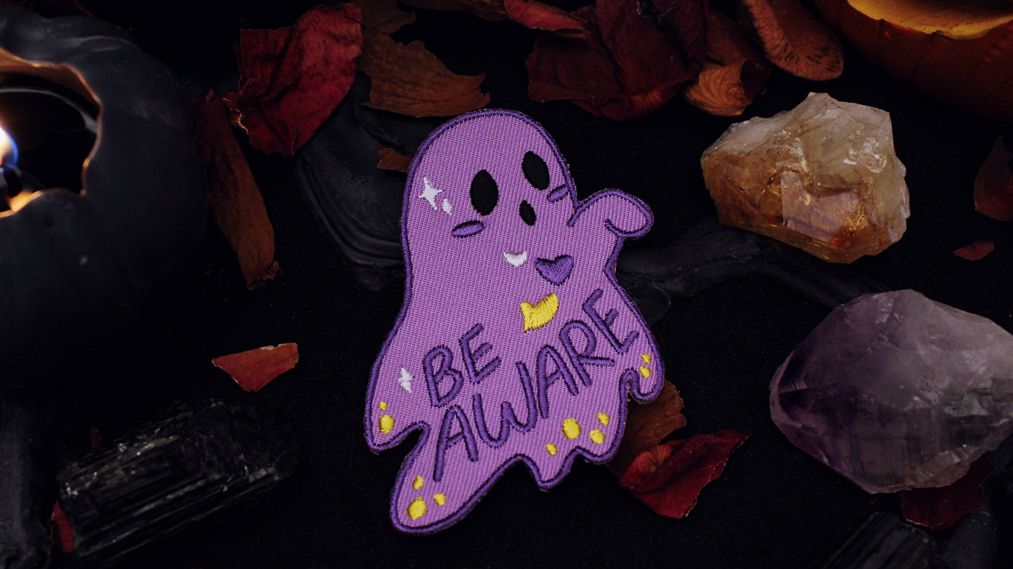 Be Aware Non-binary Patch - Iron on embroidered patch of a purple ghost with one hand sticking out and slightly bent, with the words "Be Aware" on it's body. The ghost also has black eyes and mouth, a white, purple, and yellow heart, yellow dots, and white stars, on it's body, based on the Non-binary flag colours. Original design by GRRRL Spells. 