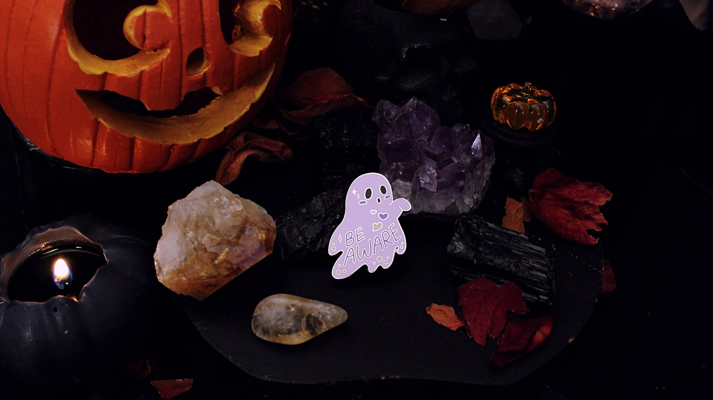 Be Aware Non-binary Pin- Silver plated enamel pin of a purple ghost with one hand sticking out and slightly bent, with the words "Be Aware" on it's body. The ghost also has black eyes and mouth, a white, purple, and yellow heart, yellow dots, and white stars, on it's body, based on the Non-binary flag colours. Original design by GRRRL Spells. 