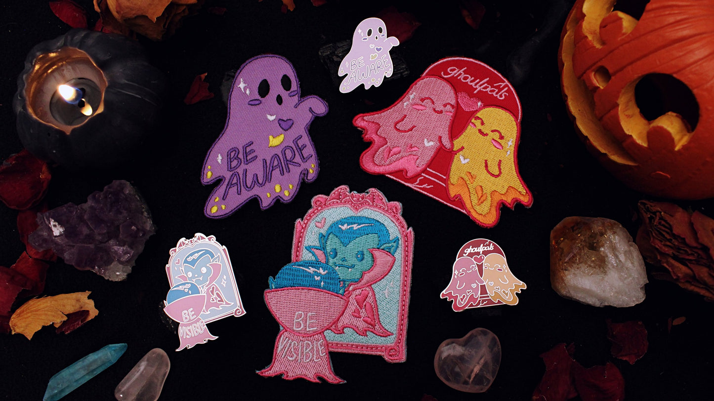 A collection of pins and patches from the HallowQueer collection, including Be Aware pin and patch, Be Visible pin and patch, and Ghoulpals pin and patch. Original design by GRRRL Spells. 