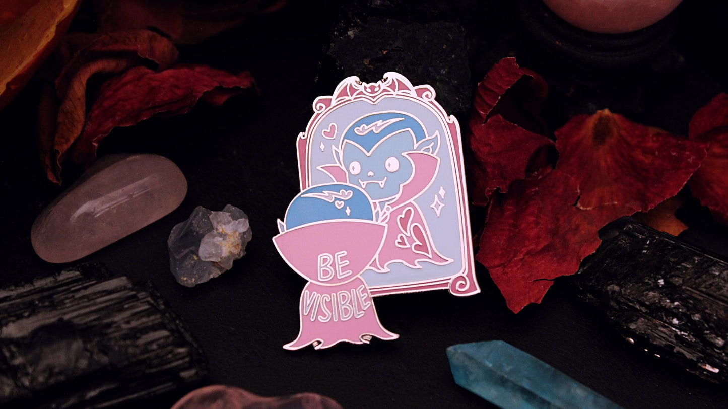 Be Visible Pin - Silver plated enamel pin of a cute vampire looking at their reflection in a mirror, in the trans flag colours of blue, pink, and white. The words Be Visible are seen on the back of their cape. Original design by GRRRL Spells. 