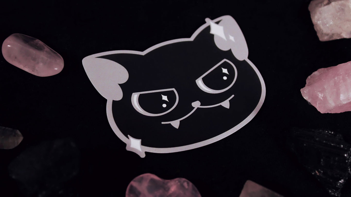 A vinyl sticker of a cute black cat with a little smirk and sparkles around its face. Original design by GRRRL Spells.