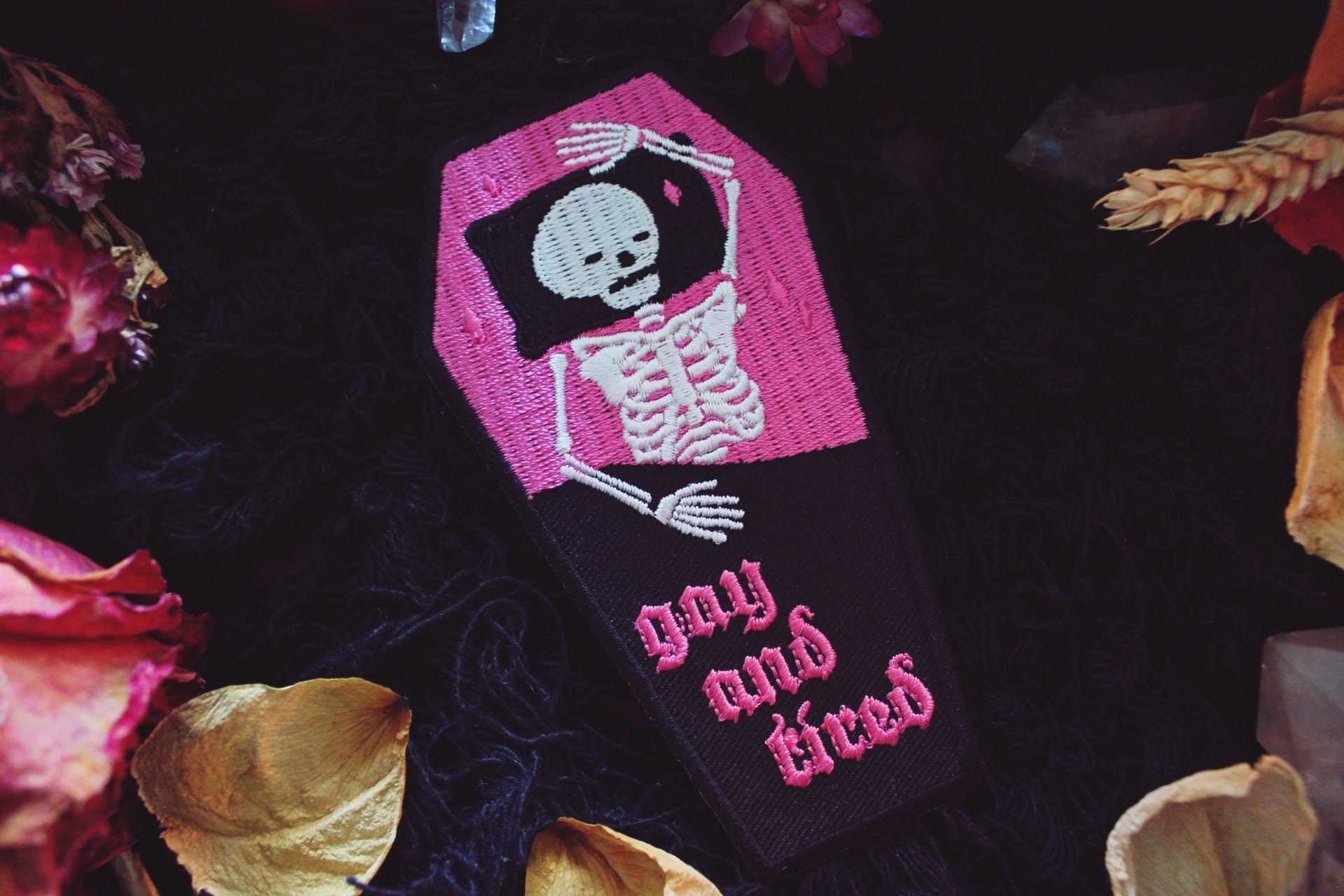 Iron on embroidered patch that is coffin shaped with black border and half pink on the top and half black on the bottom as background. Skeleton lays in the coffin bed exhausted as the words "gay and tired" in lowercase letters written on the bottom half of the coffin in gothic typography. Original design by GRRRL Spells. 