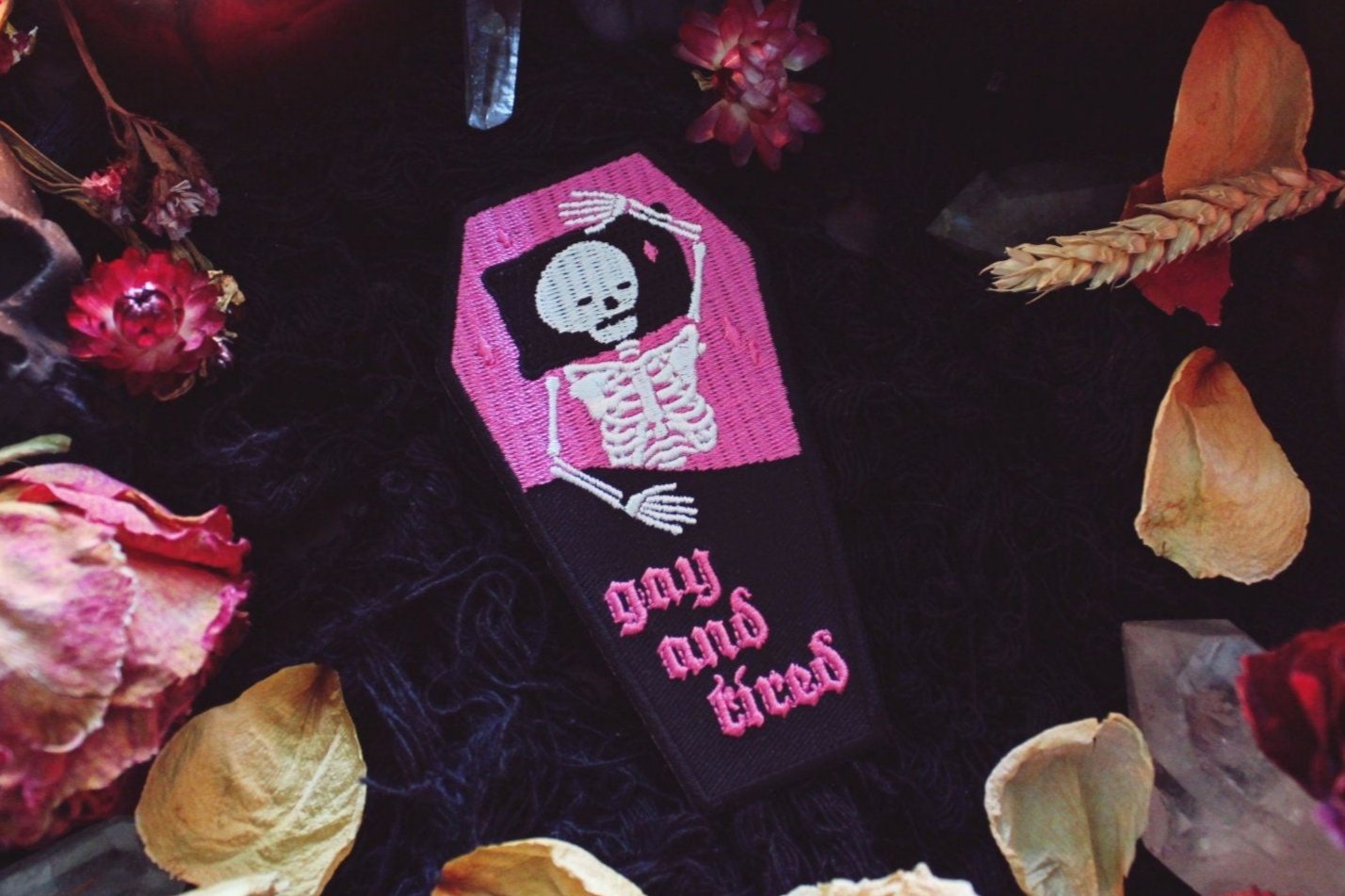 Iron on embroidered patch that is coffin shaped with black border and half pink on the top and half black on the bottom as background. Skeleton lays in the coffin bed exhausted as the words "gay and tired" in lowercase letters written on the bottom half of the coffin in gothic typography. Original design by GRRRL Spells. 