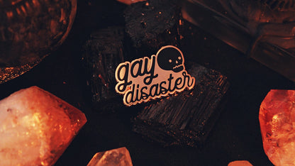 Gay Disaster Pin - Gold plated enamel pin with the words "gay disaster" written in black in lowercase in handwritten semi-cursive typography with a cute skull in the top right corner. Original design by GRRRL Spells.